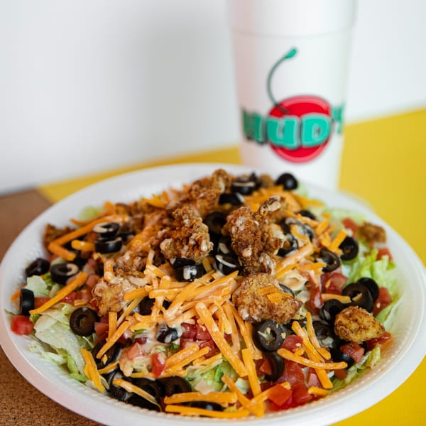 Hud's fried chicken salad with lettuce, tomatoes, olives, cheese, and dressing, with a drink cup.