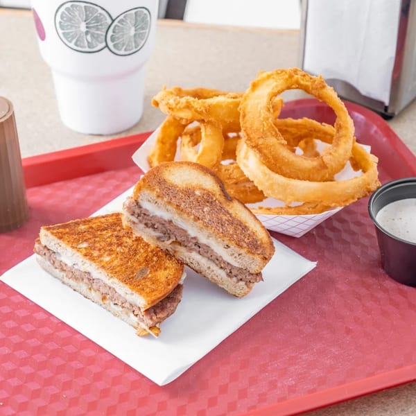 Hud's patty melt with melted cheese, crispy onion rings, and dipping sauce on a red tray.