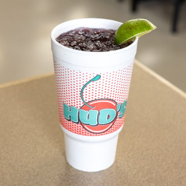 A Hud's cherry slushie in a white styrofoam cup with a lime wedge on a table.