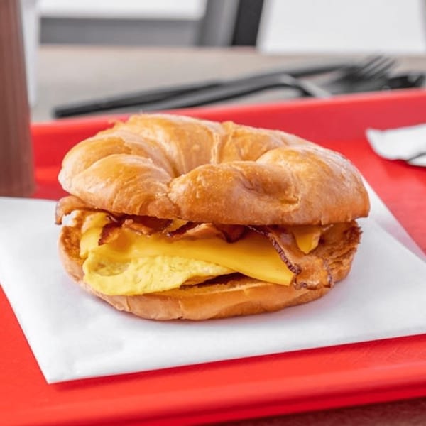A croissant breakfast sandwich with scrambled eggs, cheese, and bacon on a red tray.