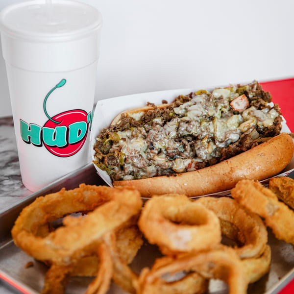 Hud's cheesesteak with onions and peppers, a drink, and onion rings on a metal tray.
