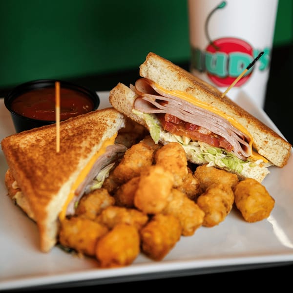 Hud's turkey club sandwich with cheese, lettuce, tomato, tater tots, and a side of sauce.