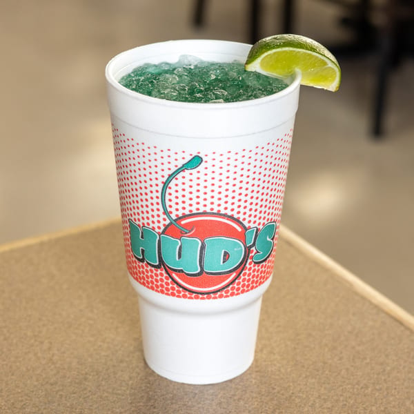 Hud's branded cup filled with a blue frozen drink, topped with ice and a lime wedge.