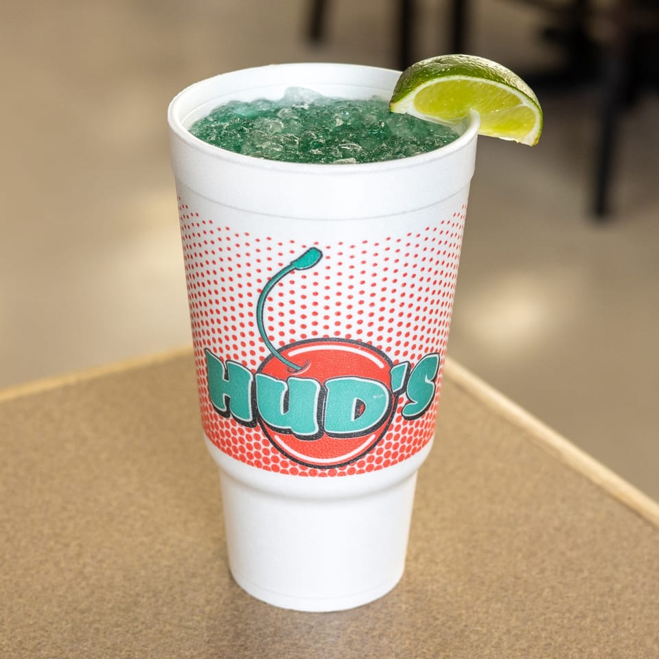 Hud's branded cup filled with a blue frozen drink, topped with ice and a lime wedge.